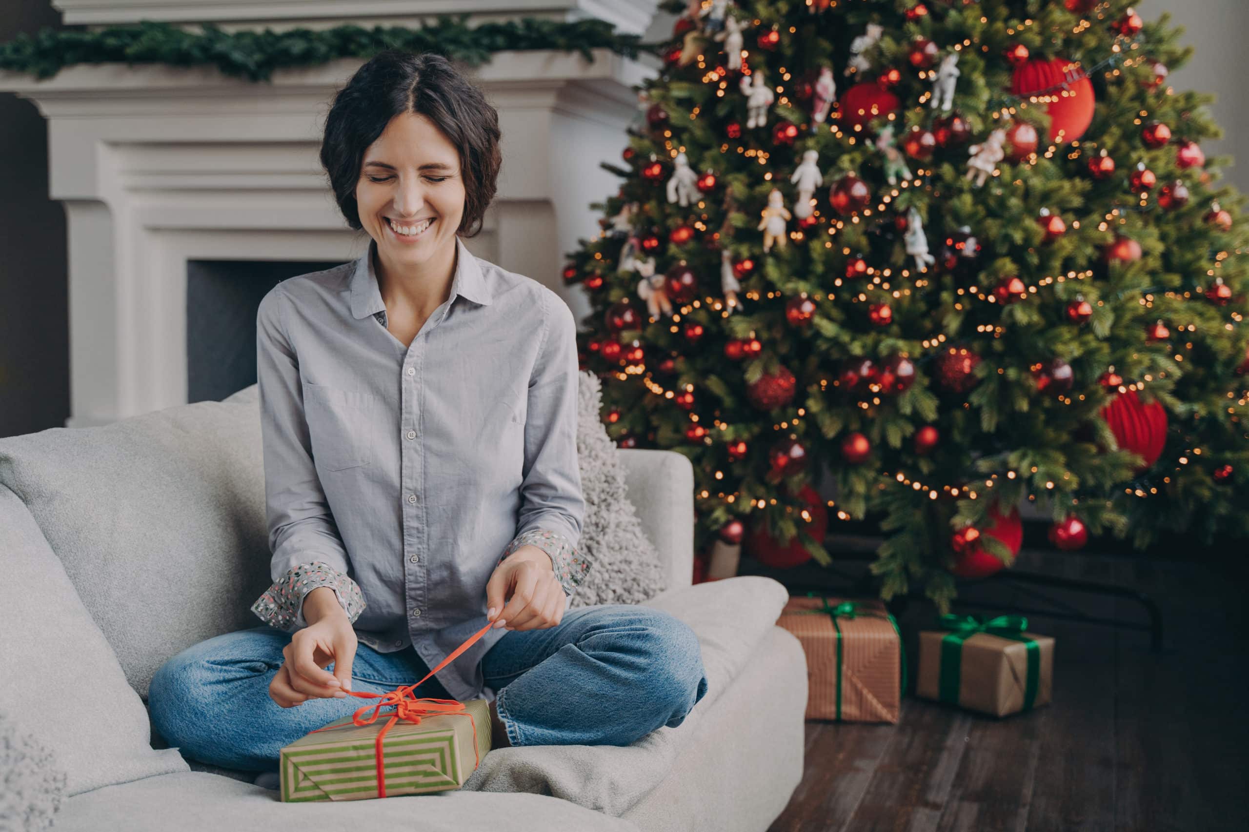 excited-italian-woman-with-closed-eyes-trying-unpack-christmas-gift-while-sitting-sofa-home-scaled